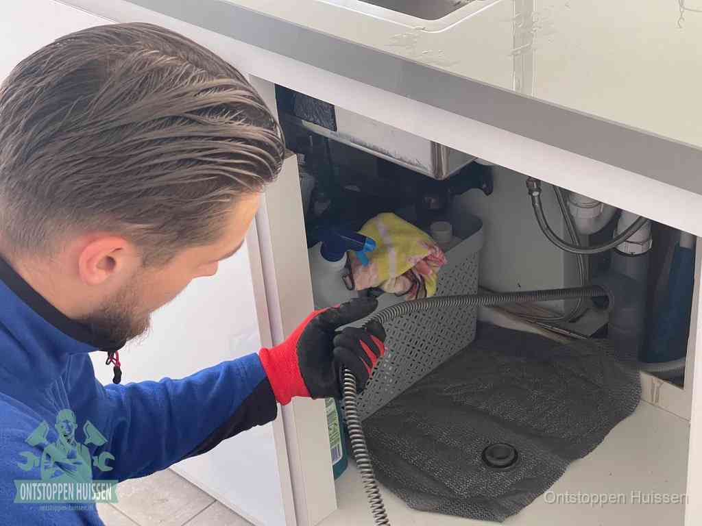 Een monteur voert een flexibele ontstoppingsveer in onder een keukenkastje om een verstopping voorbij de sifon te bereiken in Huissen