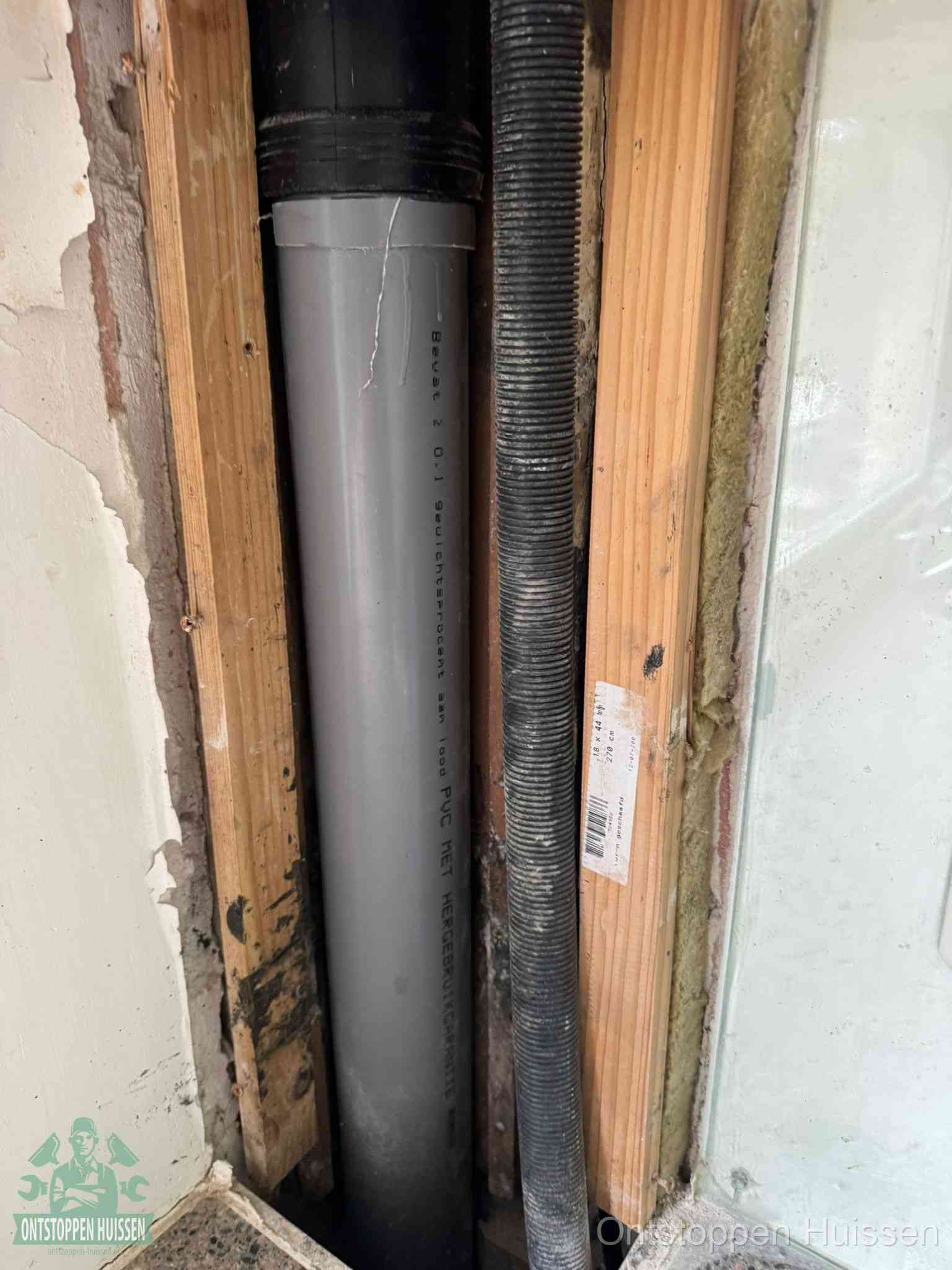 Een grijze PVC-afvoerbuis met een flexibele ribbelslang in een open wandconstructie, een verwaarloosde afvoer die te lang is blijven zitten in Huissen