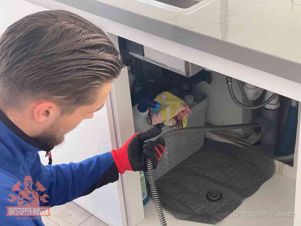 Een monteur voert een flexibele ontstoppingsveer in onder het keukenkastje, langs de sifon, om de gootsteen te ontstoppen in Huissen