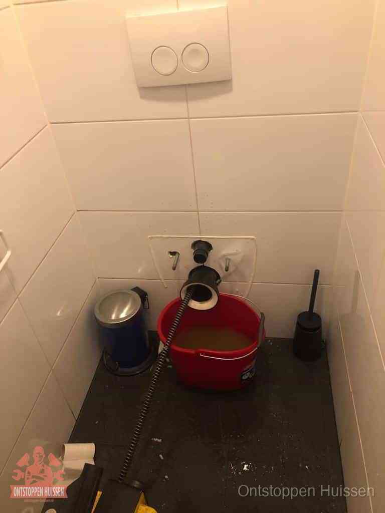 Een ontstopte wand-wc met verwijderde toiletpot waar een ontstoppingsveer in de wanduitgang verdwijnt, het achterhalen waar de verstopping zit in Huissen