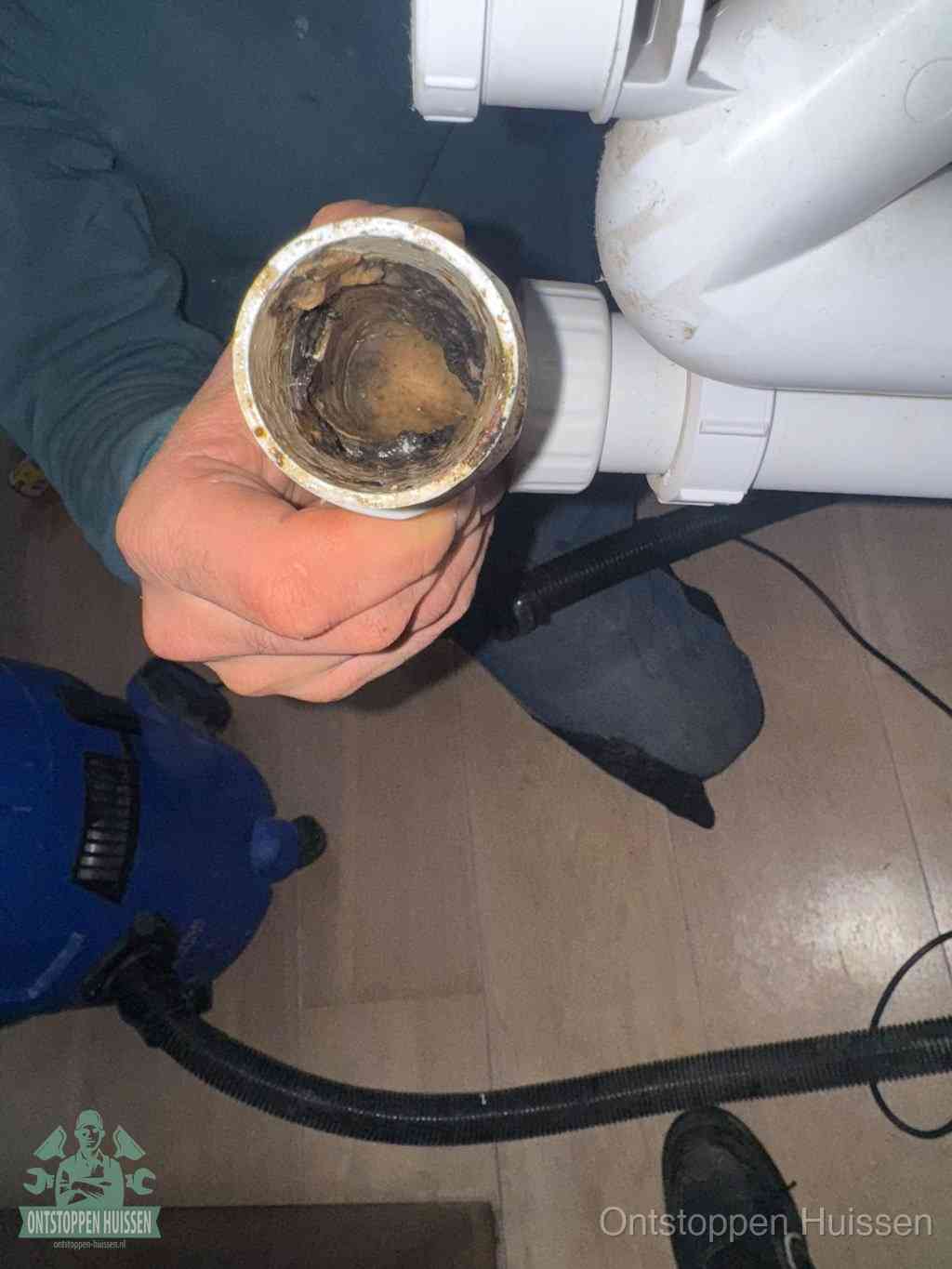 Een hand houdt een losgekoppeld PVC-afvoerbuisstuk vast dat van binnen volledig is dichtgeslibd met vet, vuil en aanslag, de meest voorkomende oorzaak van een verstopte afvoer in Huissen