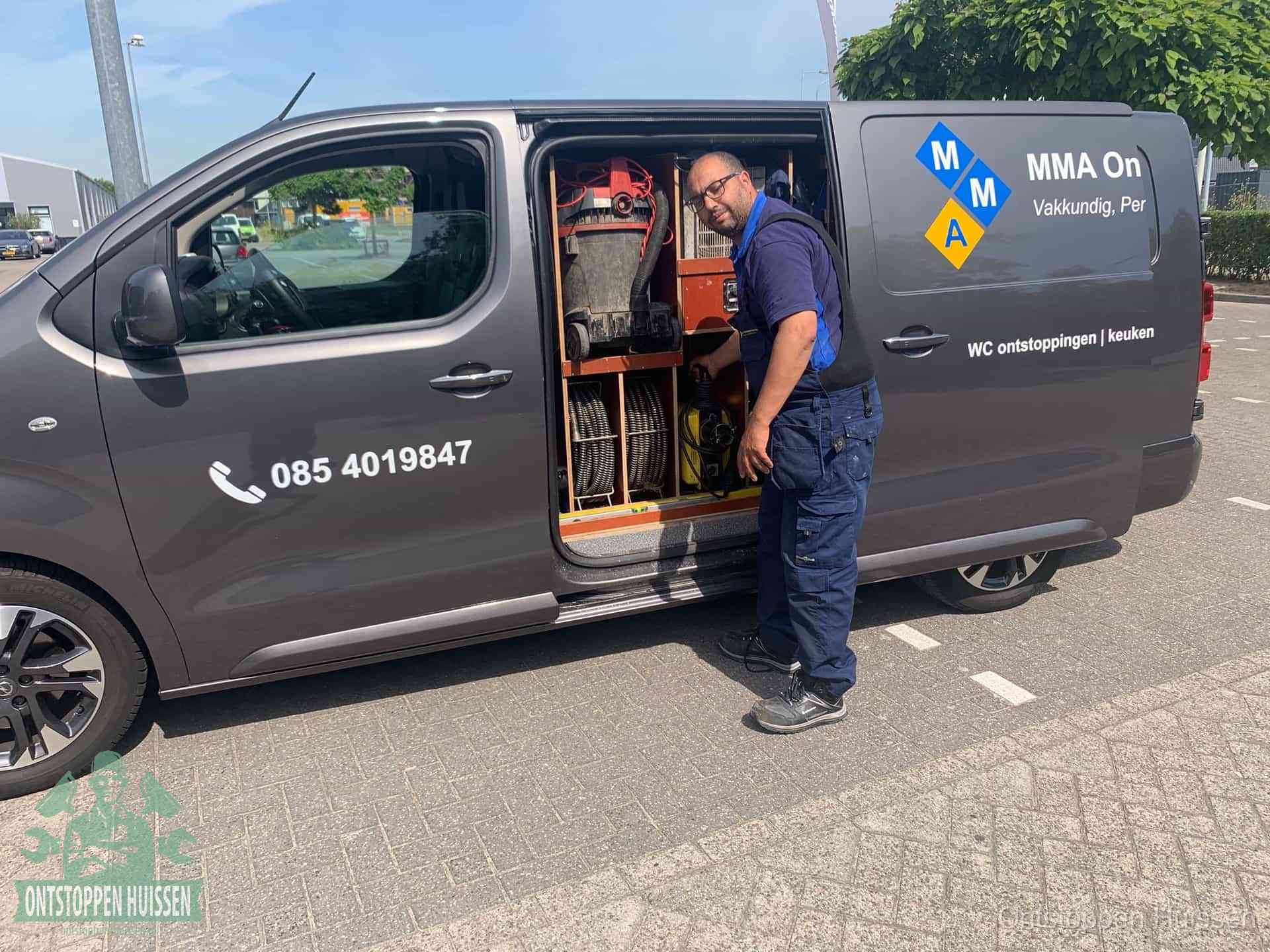 Een monteur bij de open schuifdeur van zijn bedrijfsbus op een parkeerplaats, klaar om de oorzaak van terugkerende afvoerverstoppingen aan te pakken in Huissen